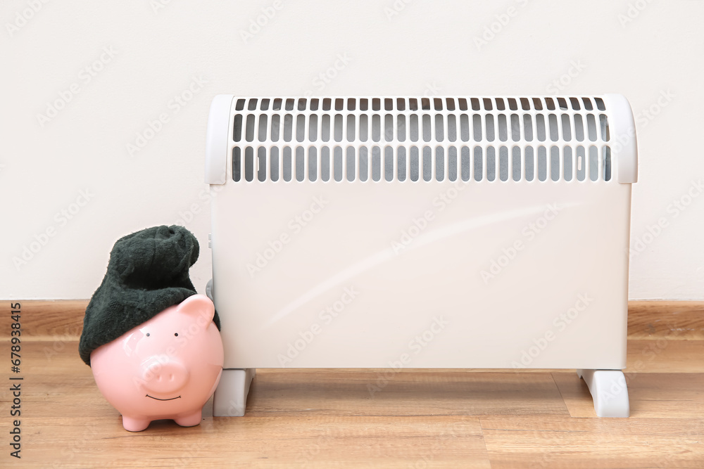 Piggy bank with warm hat near electric convector heater at home. Heating saving concept