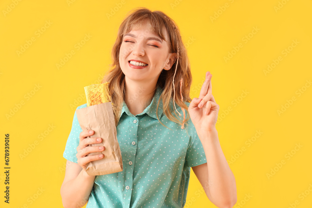 Young woman with tasty sandwich wrap crossing fingers on yellow background