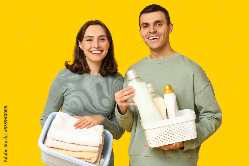 Young couple with laundry basket and bottles of detergent on yellow background