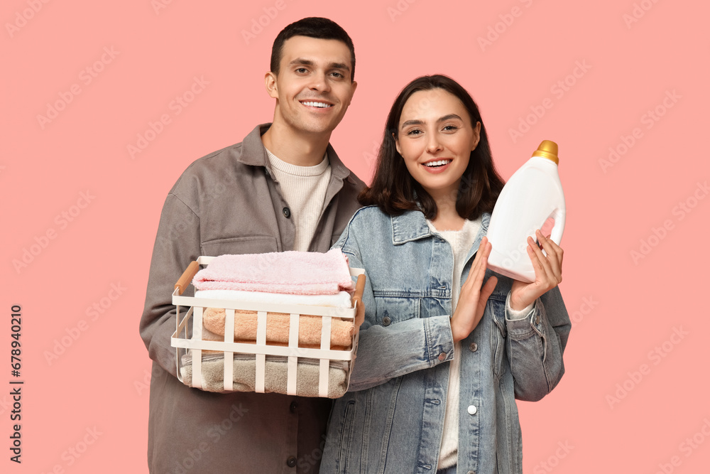 Young couple with laundry basket and bottle of detergent on pink background