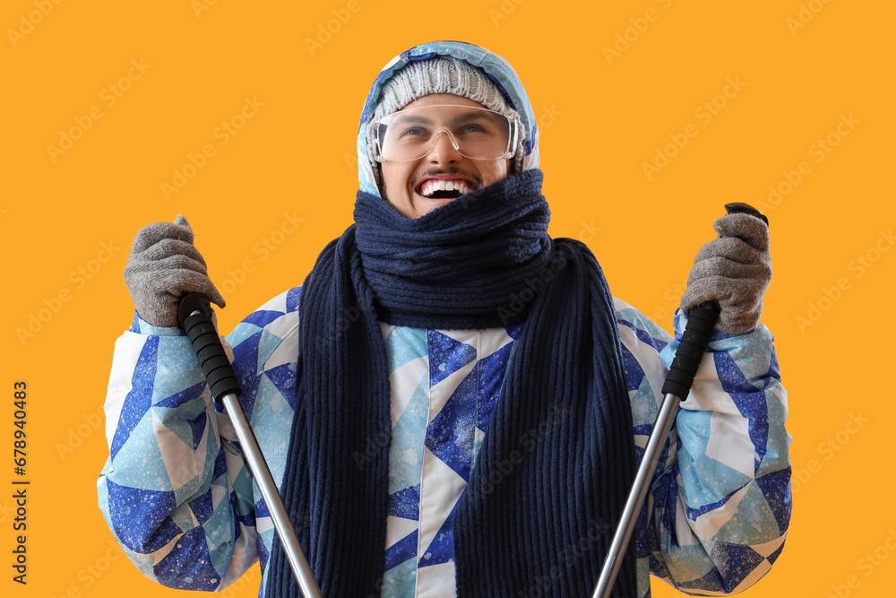 Young man in winter clothes with ski sticks on yellow background