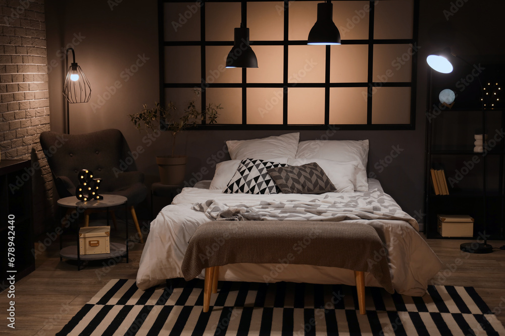 Interior of bedroom with blanket on bed and glowing lamps in evening