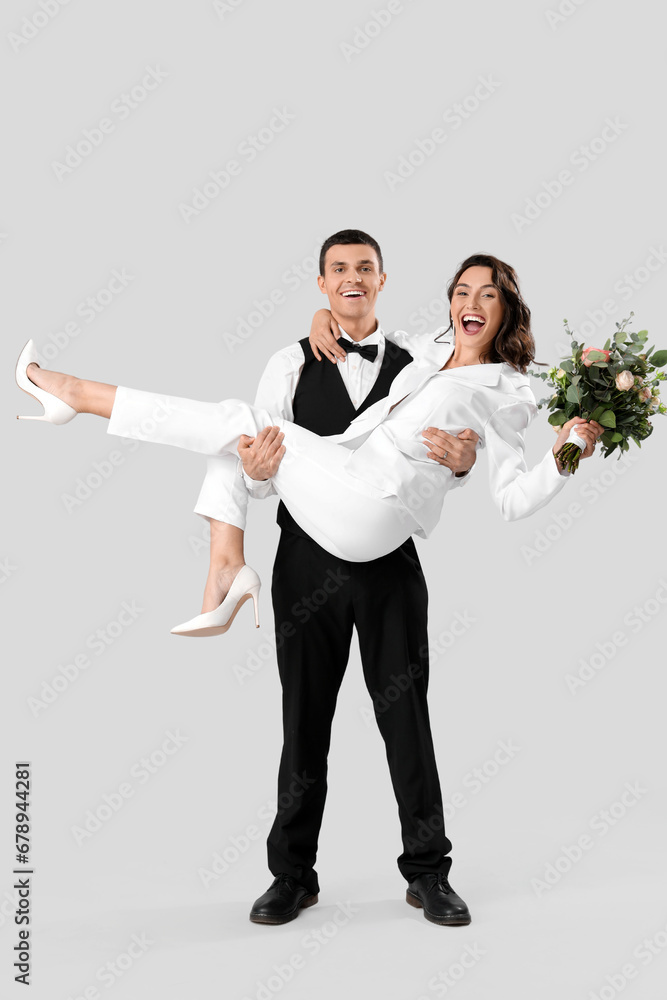 Groom holding his bride with wedding bouquet in arms on light background