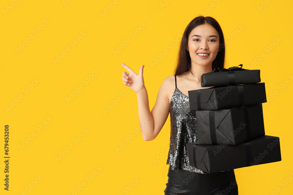 Beautiful young Asian woman with gift boxes pointing at something on yellow background. Black Friday sale