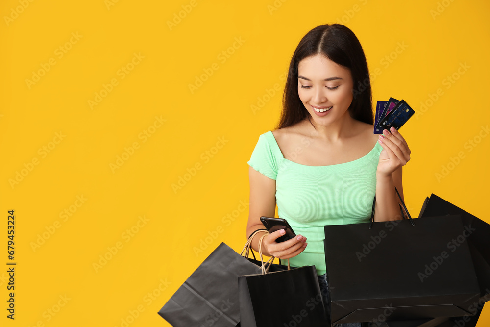 Beautiful young Asian woman with shopping bags, mobile phone and credit cards on yellow background. Black Friday sale