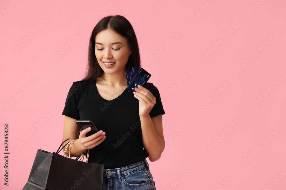 Beautiful young Asian woman with shopping bags, mobile phone and credit cards on pink background. Black Friday sale