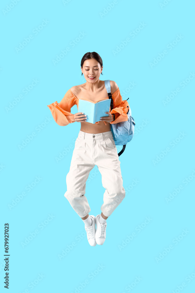 Portrait of young female student with book jumping against blue background