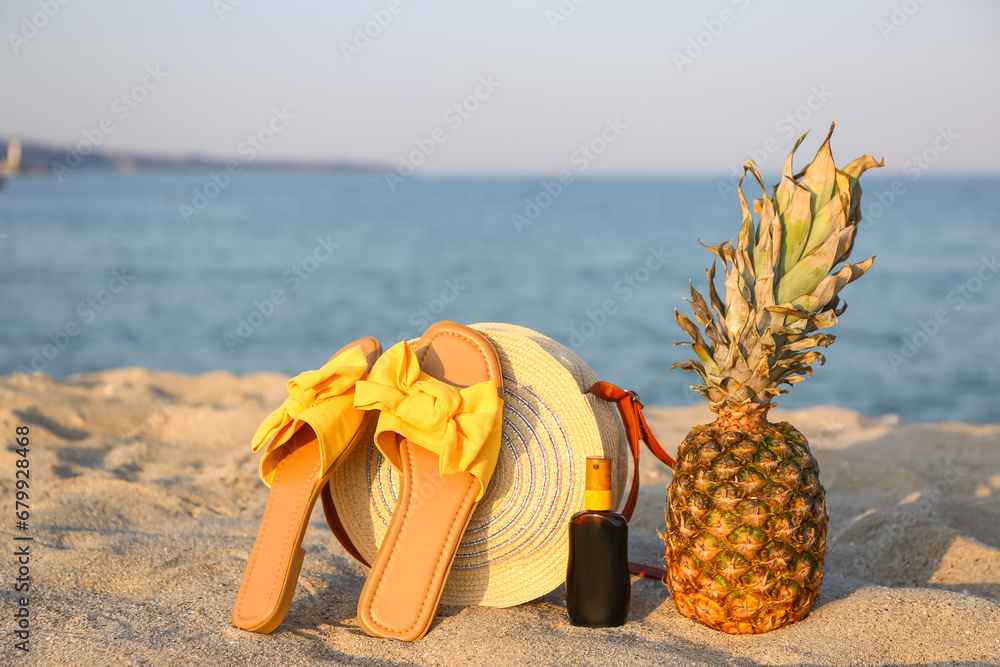 Stylish female sandals, bag, bottle of sunscreen and pineapple on sea beach