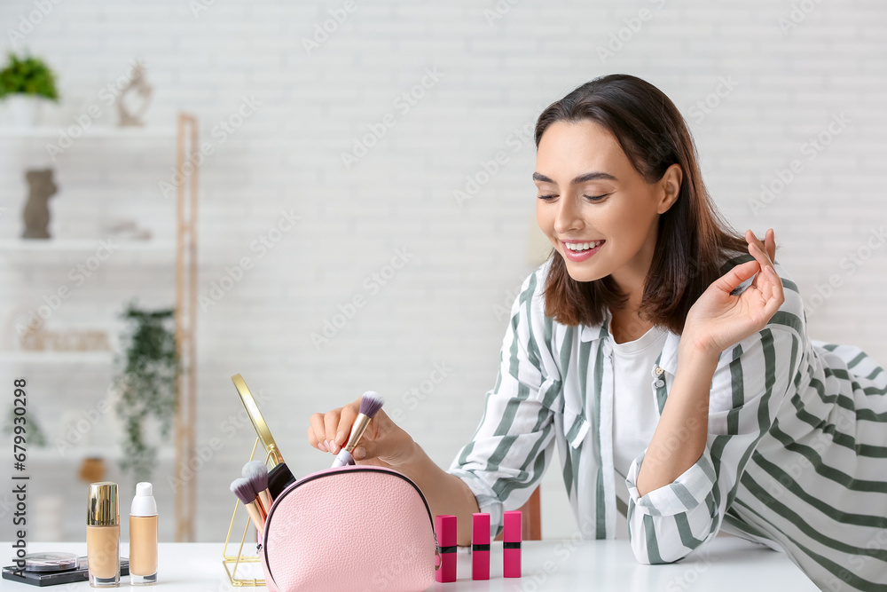 Beautiful young woman with cosmetic bag doing makeup at table in dressing room