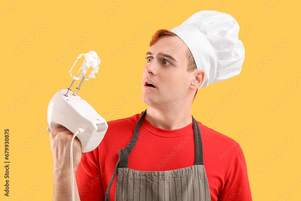 Male confectioner in apron with mixer and whipped meringue on yellow background