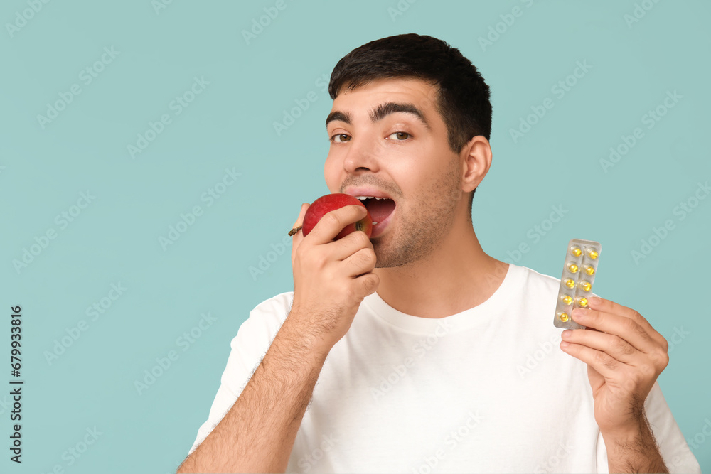 Young man with blister of vitamin A pills eating apple on blue background