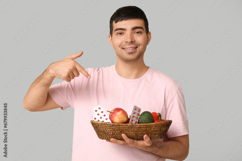 Young man pointing at basket with vitamin A pills and healthy food on light background