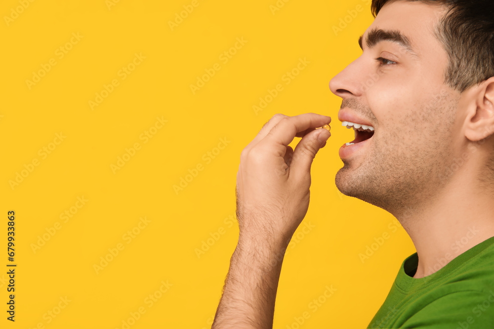 Young man taking vitamin A pill on yellow background, closeup