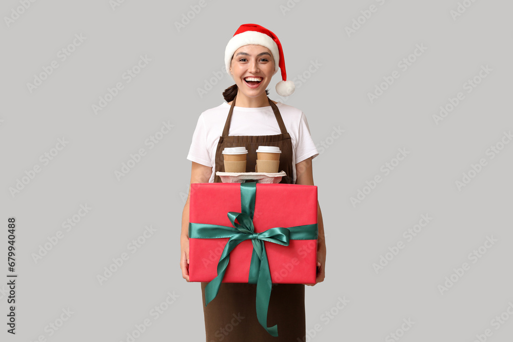 Beautiful female barista in Santa hat with cups of coffee and gift box on grey background