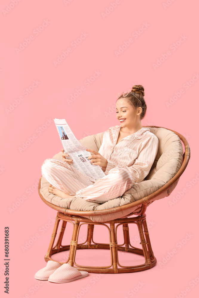 Young woman reading newspaper in armchair on pink background