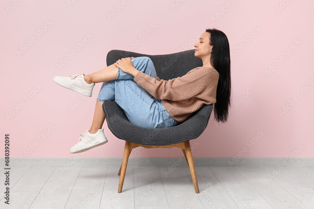 Beautiful young woman resting in comfortable armchair near pink wall