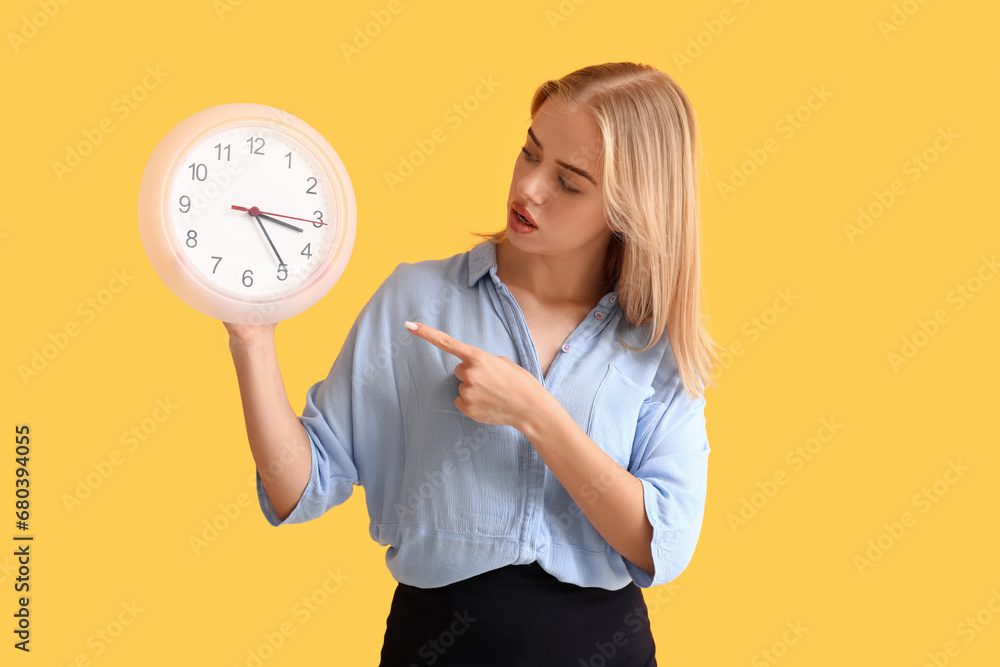 Young businesswoman pointing at clock on yellow background. Time management concept