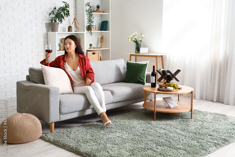 Beautiful young Asian woman with glass of wine and snacks sitting on sofa in living room