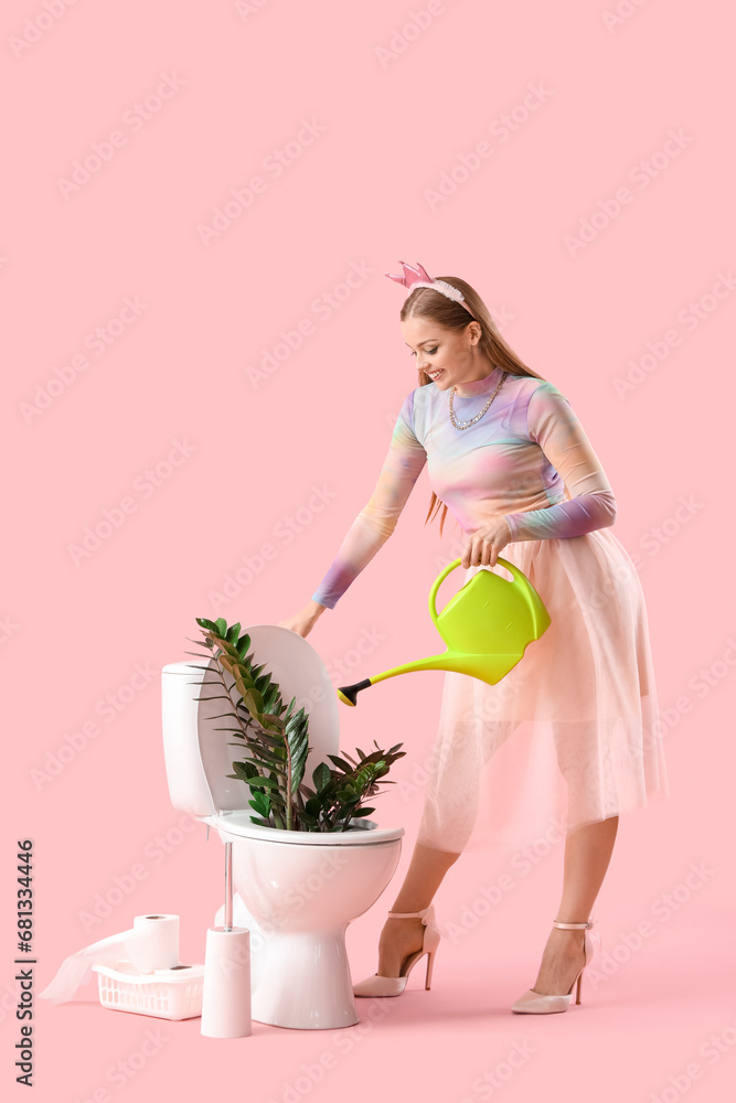 Young woman with watering can and plant in toilet bowl on pink background